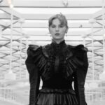 Taylor Swift in a black dress standing in the middle of a room with papers flying around it