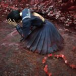 Hinako holds her head in her hand as she lays on the ground surrounded by red plants.