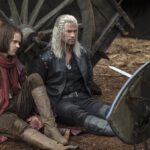 Geralt sits near a wagon of hay.