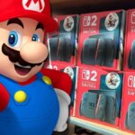 Mario gives the thumbs up in front of Switch 2s on a shelf.